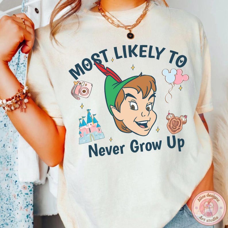 May include: Cream-colored t-shirt featuring Peter Pan with the text "Most Likely To Never Grow Up." The design includes a pink camera, a castle, balloons, and a Mickey Mouse-shaped treat. The shirt has a round neck and short sleeves.