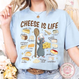 Puede incluir: Camiseta azul claro con la frase "CHEESE IS LIFE" e ilustraciones de quesos. Un chef de dibujos animados, una rata, sostiene una cuchara de madera y un trozo de queso. La camiseta presenta una variedad de nombres de quesos.