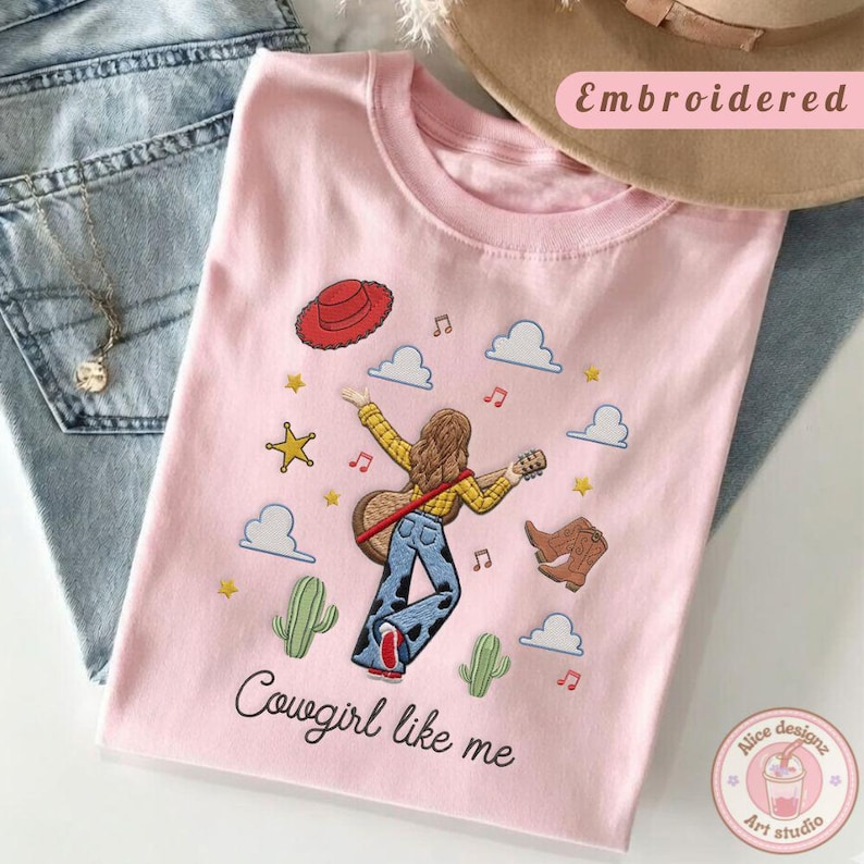 May include: A light pink t-shirt featuring an embroidered graphic of a person with long brown hair playing a guitar, wearing a yellow shirt and cow-print pants. The design includes a red hat, boots, cacti, stars, clouds, and the text Cowgirl like me.