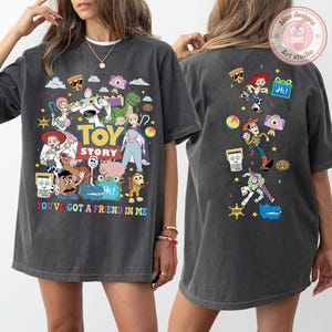 May include: A charcoal gray t-shirt featuring Toy Story character graphics on the front and back. The front displays the Toy Story logo, the text "YOU'VE GOT A FRIEND IN ME," and various characters. The back features a vertical arrangement of characters.
