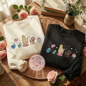 May include: Two sweatshirts, one cream and one black, with embroidered fairytale designs. The cream sweatshirt features a blonde princess, a spinning wheel, and a cake. The black sweatshirt has a similar design. Both are on a woven tray with roses.