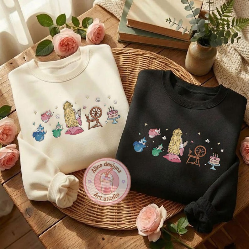 May include: Two sweatshirts, one cream and one black, with embroidered fairytale designs. The cream sweatshirt features a blonde princess, a spinning wheel, and a cake. The black sweatshirt has a similar design. Both are on a woven tray with roses.
