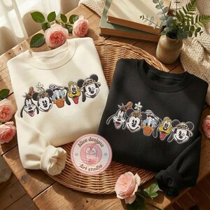 Puede incluir: Dos sudaderas de cuello redondo, una crema y otra negra, cada una con un dise&ntilde;o bordado colorido de personajes de Disney. Las sudaderas se exhiben sobre una superficie de madera con detalles florales y un posavasos decorativo.