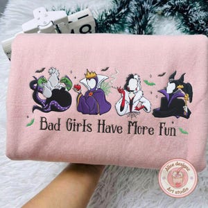 May include: A light pink sweatshirt with an embroidered design featuring four cartoon villains and the text "Bad Girls Have More Fun". The design includes bats and green accents.