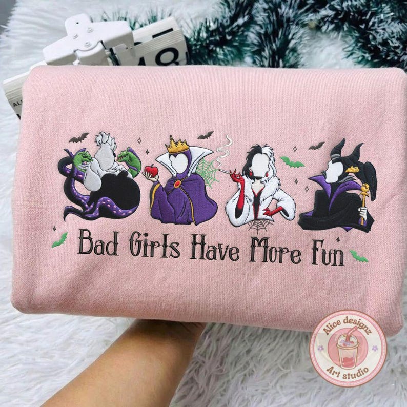 May include: A light pink sweatshirt with an embroidered design featuring four cartoon villains and the text "Bad Girls Have More Fun". The design includes bats and green accents.