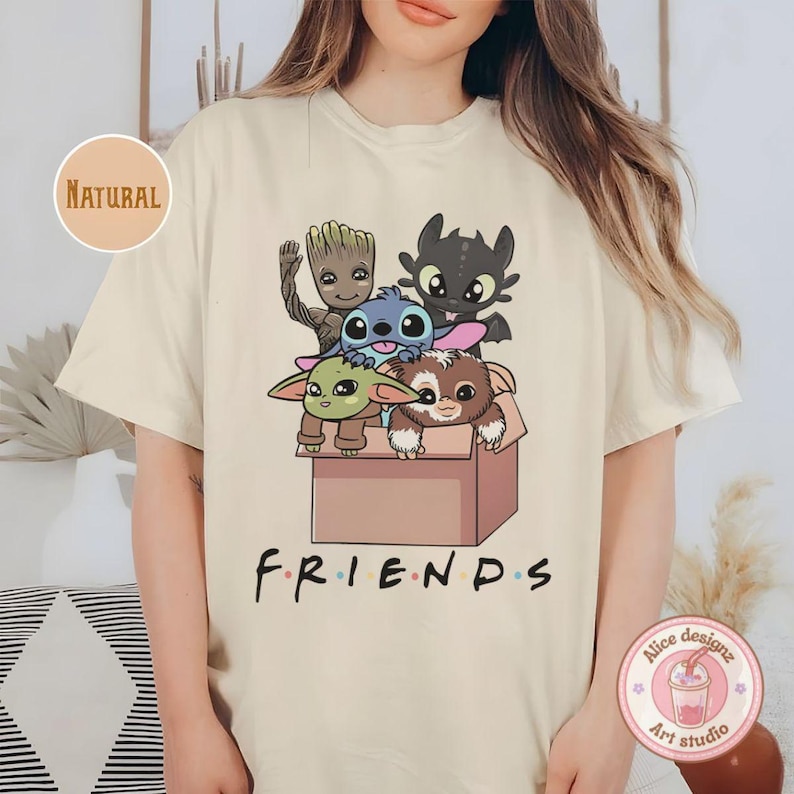 Peut inclure: T-shirt cr&egrave;me avec un motif de personnages anim&eacute;s, dont Baby Yoda, Stitch, Krokmou, Groot et un Gremlin, dans une bo&icirc;te en carton. Le mot "FRIENDS" est imprim&eacute; en noir en dessous. Le t-shirt a un col rond et des manches courtes.