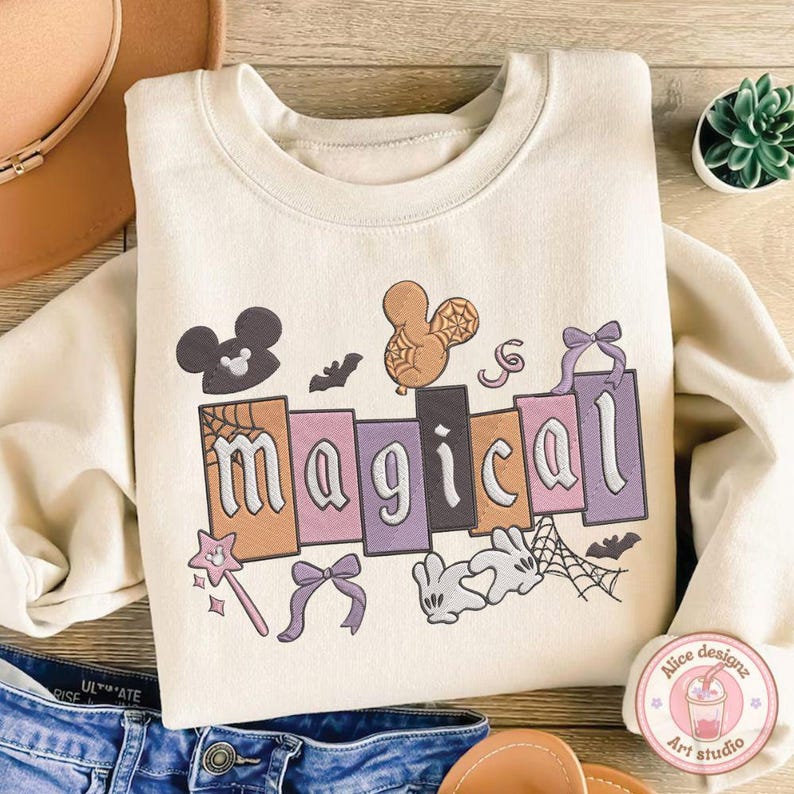 May include: A cream-coloured sweatshirt featuring the word "magical" in a block letter design. The design includes Mickey Mouse silhouettes, bats, a magic wand, and a bow. The sweatshirt is folded on top of blue jeans.