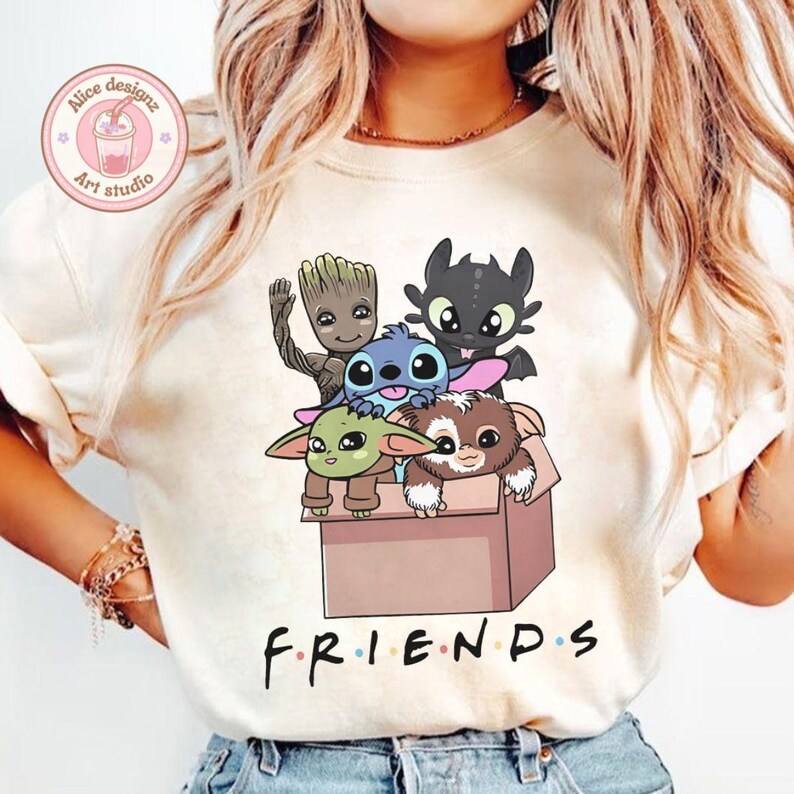Peut inclure: T-shirt cr&egrave;me avec un motif de personnages anim&eacute;s, dont Groot, Stitch, Krokmou, B&eacute;b&eacute; Yoda et Gizmo dans une bo&icirc;te en carton. Le mot "FRIENDS" est imprim&eacute; en noir en bas. Un logo est visible dans le coin sup&eacute;rieur gauche.