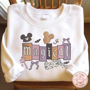 May include: White sweatshirt featuring the word "magical" in a block letter design. The design includes a Mickey Mouse head, a balloon, bats, a wand, bows, a rabbit, and a spiderweb.