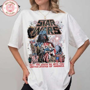 Op de afbeelding: Wit T-shirt met een Star Wars-ontwerp met Darth Vader, vuurwerk en Amerikaanse vlaggen. De tekst luidt "STAR WARS" en "MAY THE 250TH BE WITH YOU UNITED STATES OF AMERICA."