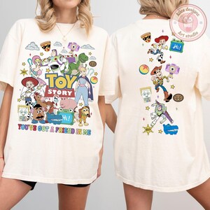 May include: An off-white t-shirt featuring Toy Story characters on the front and back. The front displays a large graphic with the text "TOY STORY" and "YOU'VE GOT A FRIEND IN ME" surrounded by various characters. The back shows a vertical arrangement of characters and icons.