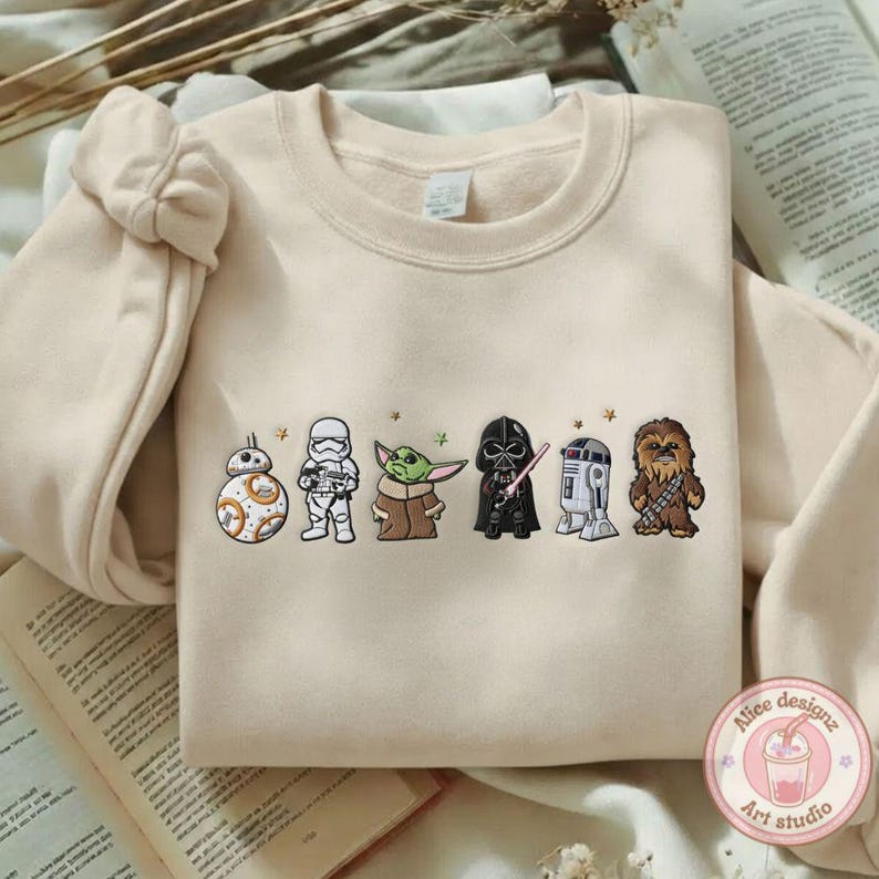 May include: A cream-colored sweatshirt featuring embroidered Star Wars characters: BB-8, a Stormtrooper, Yoda, Darth Vader, R2-D2, and Chewbacca. The design includes small star accents. The sweatshirt has a crew neck and long sleeves.