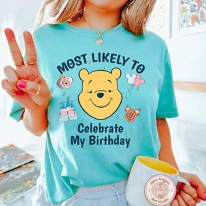 May include: A light blue t-shirt with the text "MOST LIKELY TO Celebrate My Birthday" and a graphic of Winnie the Pooh. The shirt also features graphics of a camera, castle, balloons, and a Mickey Mouse-shaped treat. A white mug with a yellow interior is also visible.