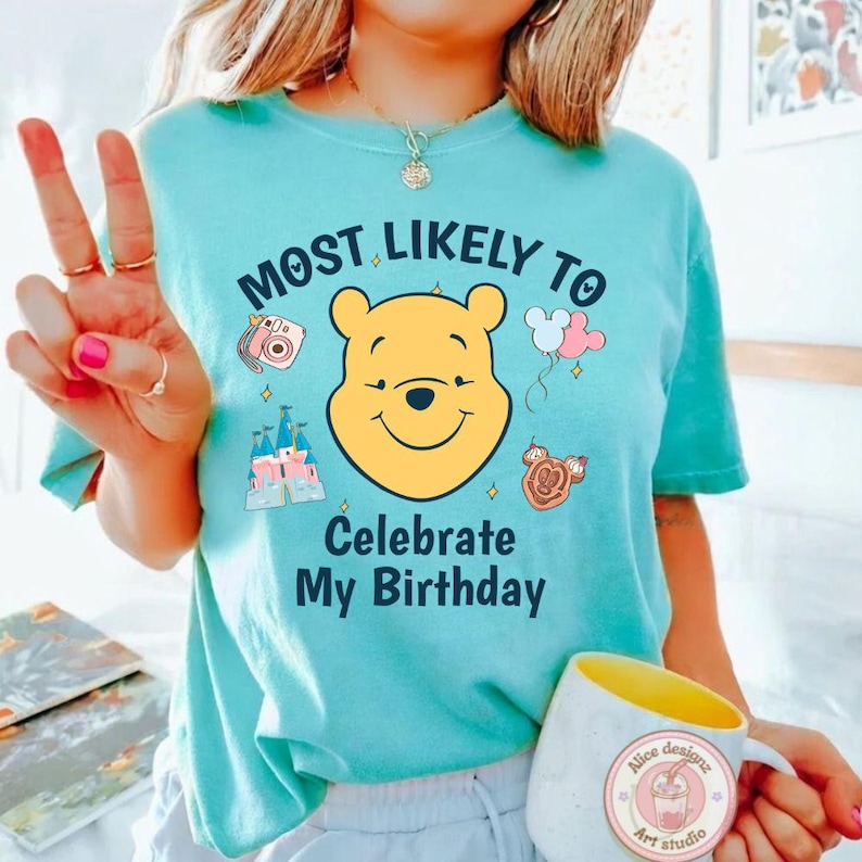 May include: A light blue t-shirt with the text "MOST LIKELY TO Celebrate My Birthday" and a graphic of Winnie the Pooh. The shirt also features graphics of a camera, castle, balloons, and a Mickey Mouse-shaped treat. A white mug with a yellow interior is also visible.