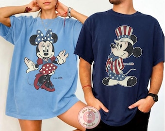 May include: Two t-shirts featuring Disney characters. One is light blue with Minnie Mouse in a red and white polka dot skirt. The other is navy blue with Mickey Mouse in patriotic attire. Both shirts have "USA 250" printed on them.