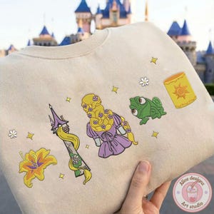 May include: Beige sweatshirt featuring embroidered images of a tower, a flower, Rapunzel, Pascal the chameleon, and a lantern. The design includes yellow, purple, green, and gold accents. The sweatshirt is held in front of a castle.