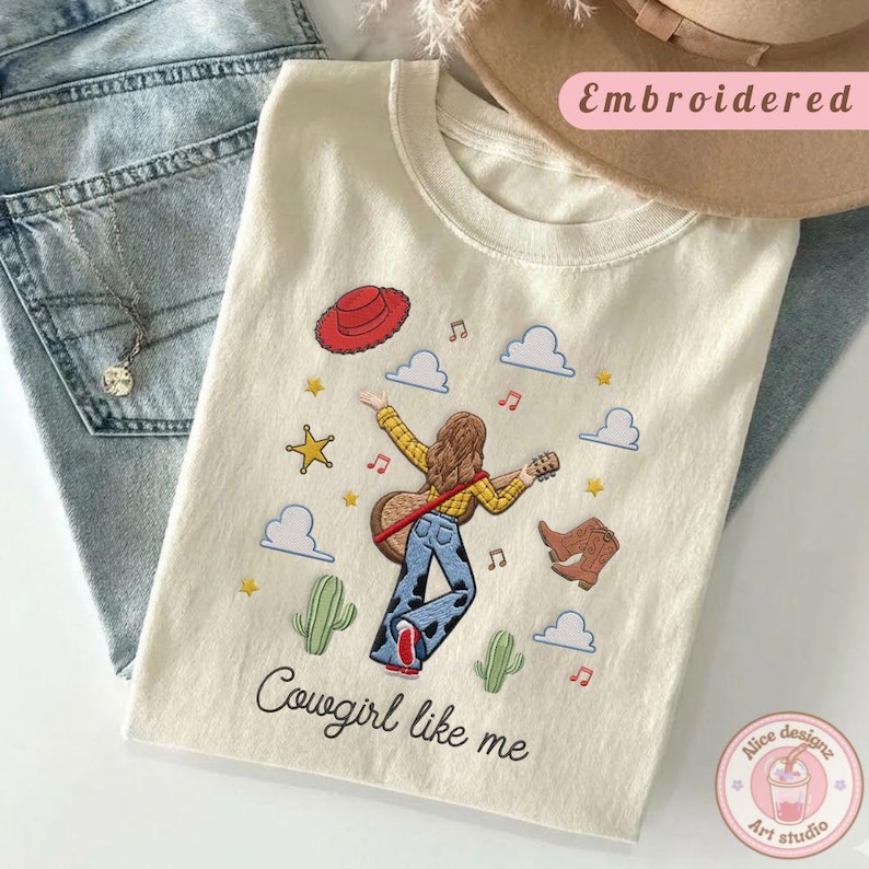 May include: A cream-colored t-shirt featuring an embroidered design of a person with long brown hair playing a guitar, wearing a yellow shirt and cow-print pants. The design includes a red hat, boots, cacti, stars, clouds, and the text Cowgirl like me.