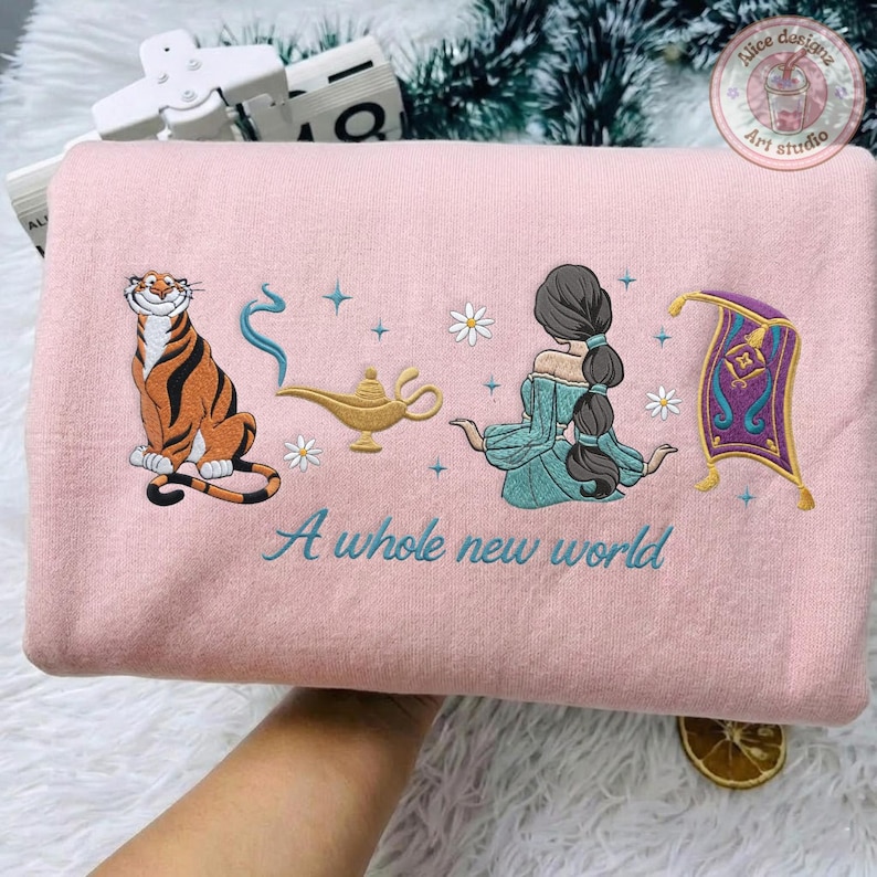 May include: A light pink sweatshirt featuring embroidered designs of a tiger, a gold lamp with blue smoke, a person with long dark hair in a teal outfit, and a purple magic carpet. The text A whole new world is embroidered in teal script.