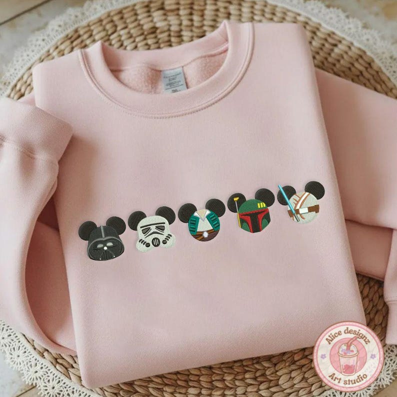 May include: A light pink sweatshirt featuring embroidered character designs. The designs include Darth Vader, a Stormtrooper, and other characters. The sweatshirt has a crew neck and long sleeves. The design is in the style of Mickey Mouse.