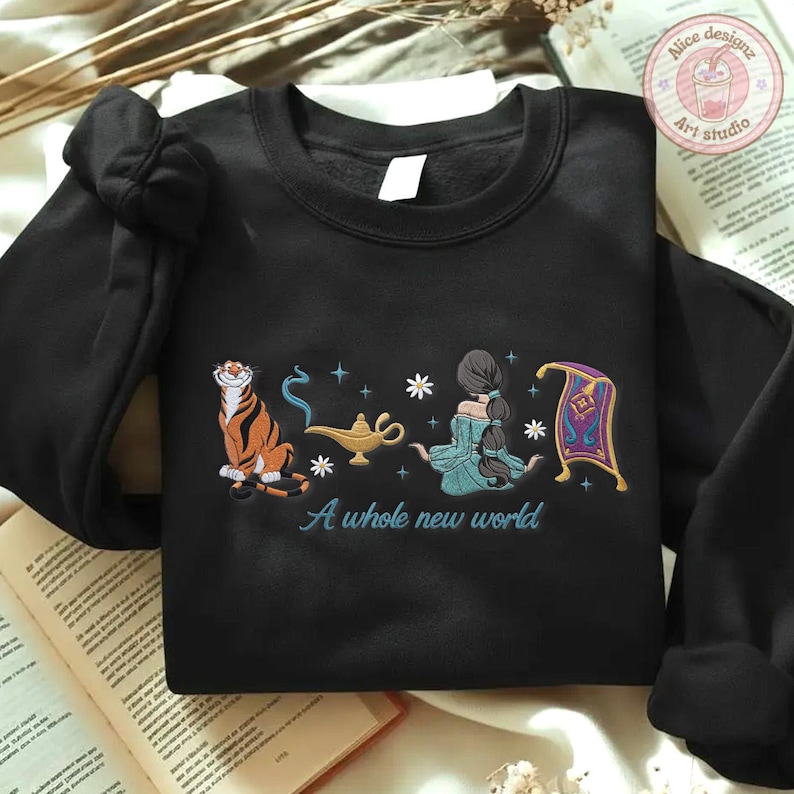 May include: A black crewneck sweatshirt featuring an embroidered design of a tiger, a gold magic lamp with blue smoke, a person with long dark hair in a teal outfit, and a purple magic carpet, with the text A whole new world in blue script.
