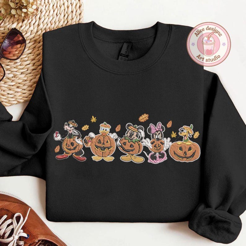 Puede incluir: Sudadera negra de cuello redondo con un dise&ntilde;o de personajes de dibujos animados disfrazados de calabazas, incluyendo Mickey y Minnie Mouse, Donald Duck y Goofy. El dise&ntilde;o incluye hojas de oto&ntilde;o y el texto "Alice designz Art studio".