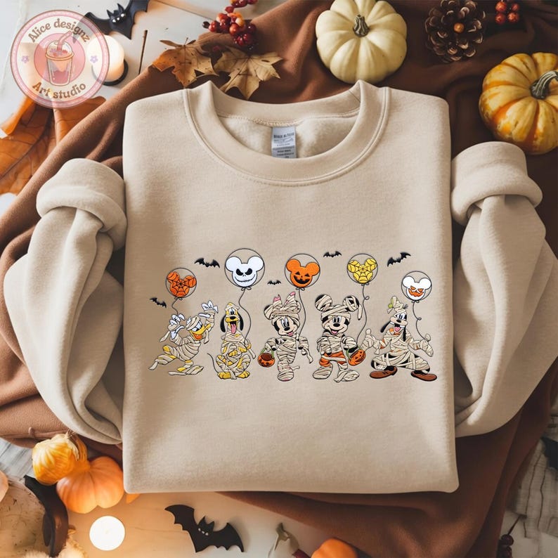 May include: Beige crewneck sweatshirt featuring a Halloween-themed graphic. The graphic depicts cartoon characters dressed as mummies, holding balloon-shaped heads. The balloons are orange, white, and yellow. Small black bats are scattered around the balloons.