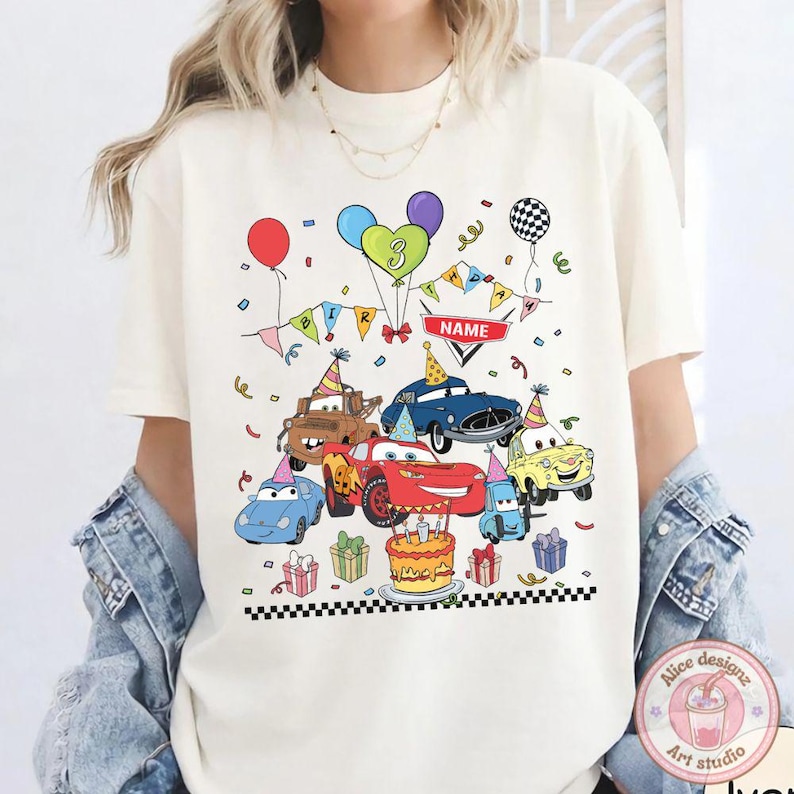 Op de afbeelding: Cr&egrave;mekleurig T-shirt met een kleurrijke afbeelding van tekenfilmauto's, ballonnen, confetti en een verjaardagstaart. Het ontwerp bevat het nummer 3 en een banner met de tekst "NAME". Een feestelijk ontwerp voor een verjaardag.