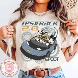 May include: Beige t-shirt featuring a graphic of Mickey Mouse, Donald Duck, and Goofy in a blue car with the text "Test Track 2.0" and "Epcot". The design includes gear illustrations.