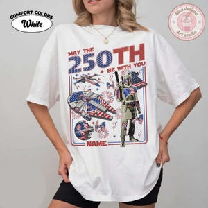 May include: White Comfort Colors t-shirt with a patriotic design featuring a spaceship, a character, and the text "MAY THE 250TH BE WITH YOU". The design incorporates red, white, and blue colours, and the text "NAME" is also visible.