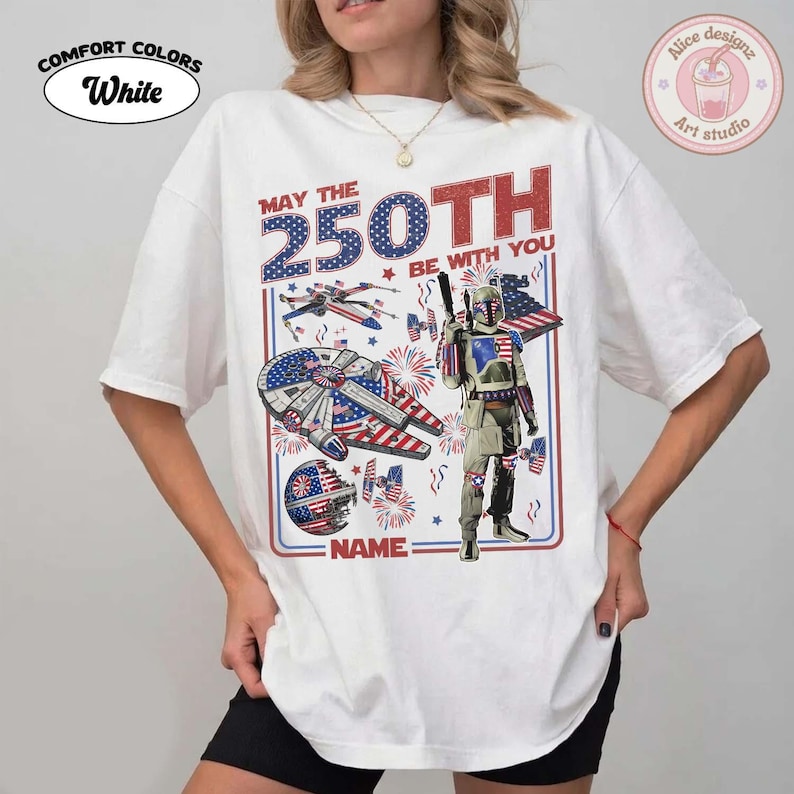 May include: White Comfort Colors t-shirt with a patriotic design featuring a spaceship, a character, and the text "MAY THE 250TH BE WITH YOU". The design incorporates red, white, and blue colours, and the text "NAME" is also visible.