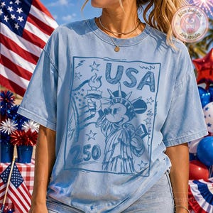 May include: Light blue t-shirt featuring a graphic of Mickey Mouse as the Statue of Liberty, holding a torch and a scroll that says "July". The word "USA" is above the image, with the number "250" below. The shirt has a vintage, faded look.