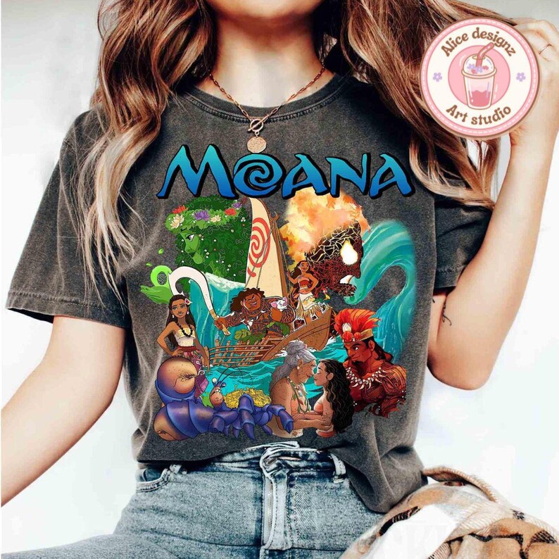 Puede incluir: Una camiseta gris con un estampado gr&aacute;fico de personajes de la pel&iacute;cula animada Moana. El gr&aacute;fico incluye a Moana, Maui y otros personajes de la pel&iacute;cula. El texto "Moana" est&aacute; impreso en letras grandes en la camiseta.