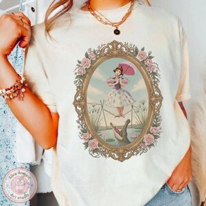May include: A cream-colored t-shirt featuring a graphic print of a person in a dress holding a pink umbrella while balancing on a tightrope above a crocodile, all enclosed within an ornate gold oval frame decorated with pink roses.