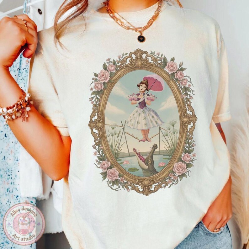 May include: A cream-colored t-shirt featuring a graphic print of a person in a dress holding a pink umbrella while balancing on a tightrope above a crocodile, all enclosed within an ornate gold oval frame decorated with pink roses.