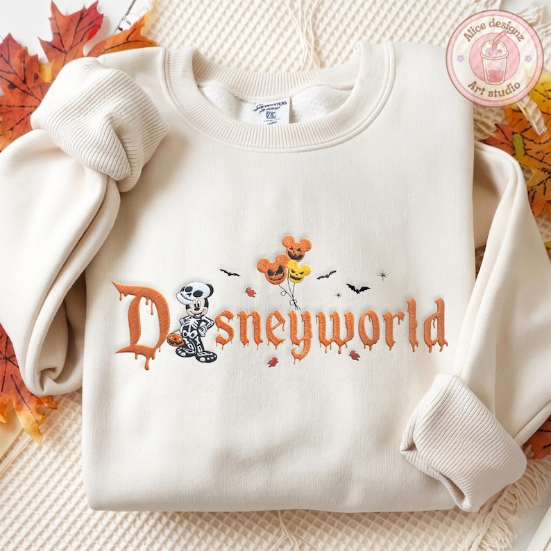 May include: A cream-colored crewneck sweatshirt featuring embroidered text that reads Disneyworld in an orange dripping font. The design includes a skeleton-costumed mouse character, three pumpkin-shaped balloons, and small black bats.