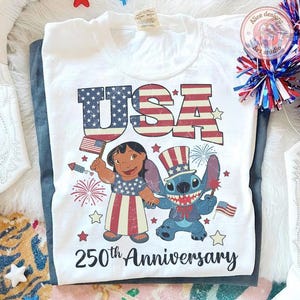 May include: A white t-shirt featuring a graphic of Lilo and Stitch dressed in patriotic attire with the text USA and 250th Anniversary. The design includes red, white, and blue stars, stripes, fireworks, and small American flags in a distressed style.