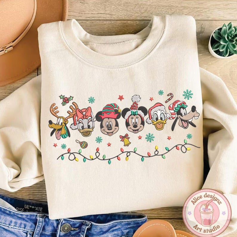 Op de afbeelding: Cr&egrave;mekleurige sweatshirt met geborduurde cartoonfiguren in feestelijke kerstoutfit. Het ontwerp omvat Goofy, Donald Duck, Mickey Mouse, Minnie Mouse en andere personages, met kerstverlichting en sneeuwvlokken.