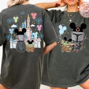 May include: Dark gray t-shirts featuring a cartoon illustration of The Mandalorian and Grogu with Mickey Mouse ears, in front of a castle and balloons. The design includes fireworks and a castle.