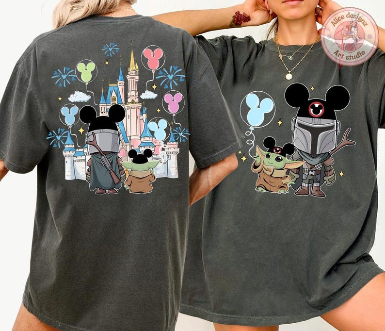 May include: Dark gray t-shirts featuring a cartoon illustration of The Mandalorian and Grogu with Mickey Mouse ears, in front of a castle and balloons. The design includes fireworks and a castle.