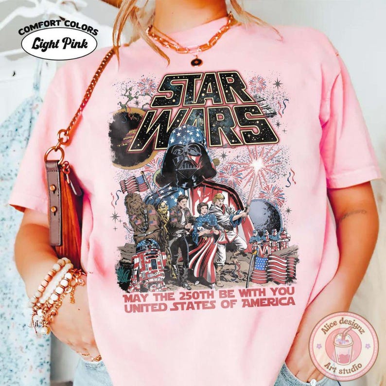 Op de afbeelding: Lichtroze Comfort Colors T-shirt met een Star Wars-ontwerp, inclusief Darth Vader, vuurwerk en de tekst "MAY THE 250TH BE WITH YOU UNITED STATES OF AMERICA." Het shirt heeft ook de tekst "Alice designz Art studio."