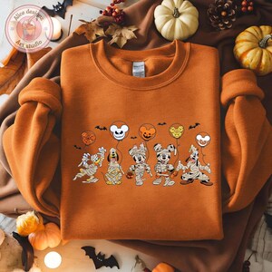 May include: Orange sweatshirt featuring a Halloween-themed graphic of cartoon characters dressed as mummies, holding balloon-shaped heads. The balloons are orange, yellow, and white. Black bats are scattered around the design. The sweatshirt has a crew neck.