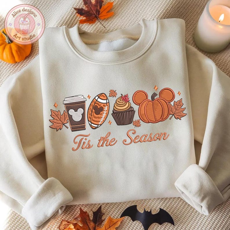 May include: Cream-colored sweatshirt with an embroidered fall-themed design. The design includes a coffee cup, football, cupcake, and pumpkin, all with Mickey Mouse-inspired details. The text "Tis the Season" is below the design. A small pumpkin and leaves are visible.