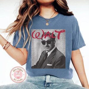 Op de afbeelding: Blauw T-shirt met een zwart-wit afbeelding van Walt Disney met een zonnebril en het woord "WALT" in rode letters. Het shirt heeft een ronde hals en korte mouwen.