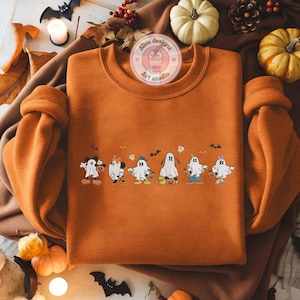 K&ouml;nnte beinhalten: Orangenes Sweatshirt mit Rundhalsausschnitt und einer Reihe von Cartoon-Geistern, die jeweils einen K&uuml;rbis halten. Das Design umfasst Flederm&auml;use und kleine Geister. Das Sweatshirt wird auf einer braunen Oberfl&auml;che mit K&uuml;rbissen und Herbstdekorationen pr&auml;sentiert.
