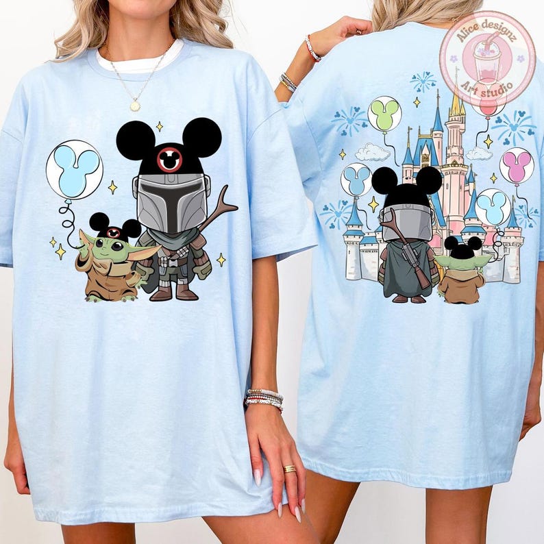 May include: Light blue t-shirt featuring a cartoon illustration of The Mandalorian and Grogu with Mickey Mouse ears. The back of the shirt shows the characters in front of a castle with balloons. The shirt has the text "Alice designz Art studio".