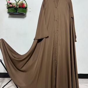 Puede incluir: Un vestido abaya largo y fluido de color marrón con botones en la parte delantera y mangas anchas. El vestido se exhibe en una percha, mostrando su elegante caída. Un arreglo floral decorativo es visible en el fondo.