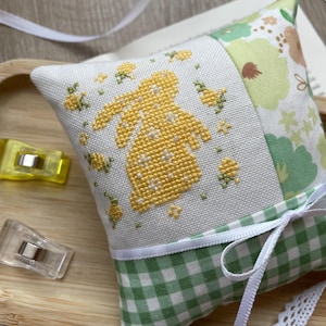 May include: A square pin cushion with a cross-stitched yellow bunny design on white fabric. The cushion features green and white gingham and floral patterned fabric, and is tied with a white ribbon. Two yellow clips are visible.