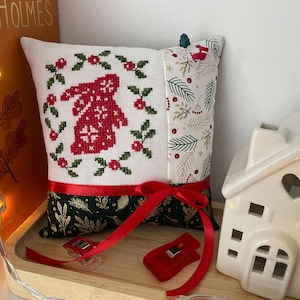 May include: A decorative Christmas pillow with a red embroidered bunny design, surrounded by green floral accents. The pillow features a red ribbon and a patterned fabric with green leaves and berries. A small white ceramic house is in the background.