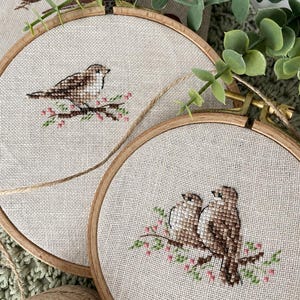 Könnte beinhalten: Zwei runde Stickrahmen aus Holz mit Kreuzstichmotiven auf hellbeigem Stoff. Ein Rahmen zeigt einen einzelnen braunen Vogel auf einem Zweig mit rosa Blüten, der andere zwei braune Vögel, die auf einem Zweig mit rosa Blüten sitzen.