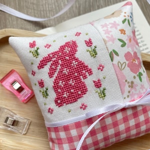 May include: A handmade pincushion with a cross-stitched pink bunny design on white fabric. The pincushion features floral and gingham patterns, with a white ribbon accent. Green floral accents and pink tulips surround the bunny.